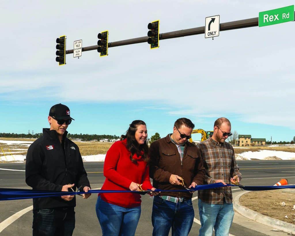 Four people stand holding scissors, cutting a blue ribbon at a road intersection with traffic lights and a street sign labeled "Rex Rd.