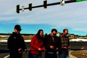 Four people stand holding scissors, cutting a blue ribbon at a road intersection with traffic lights and a street sign labeled "Rex Rd.