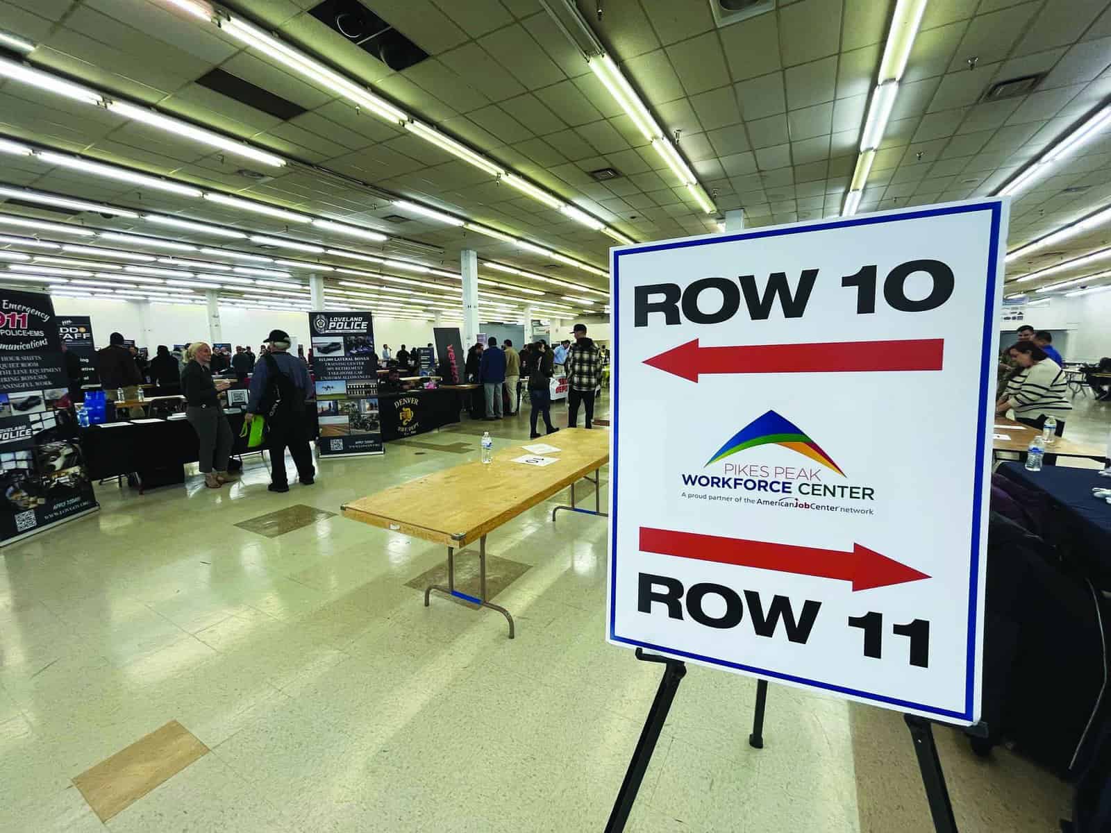 Job fair with booths set up in a large room. A sign points to Rows 10 and 11, labeled "Pikes Peak Workforce Center." People are visible in the background exploring the event.