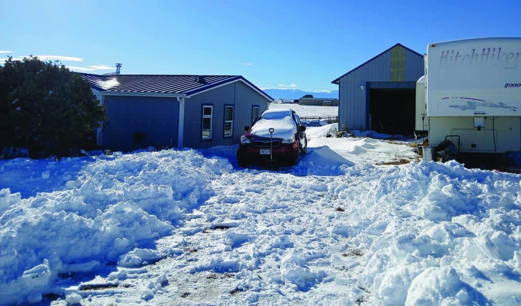 A snow-covered driveway with a car, house, and garage in a rural setting. Snow is piled on the ground and the roof of the car. A trailer is parked nearby.