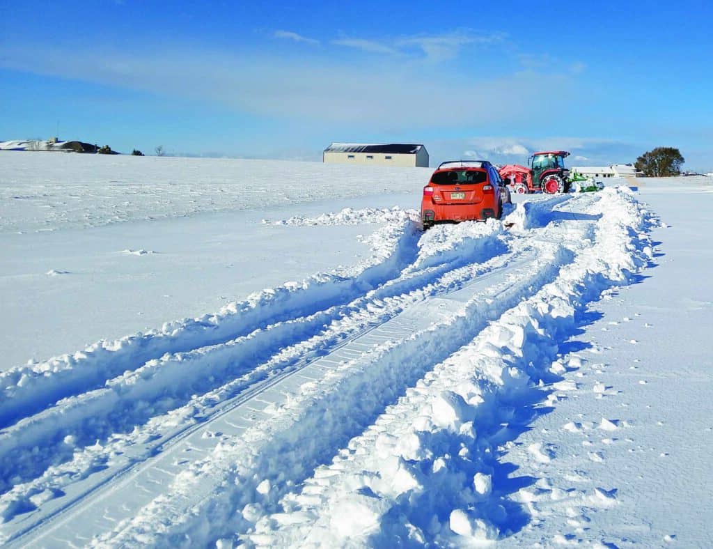 A tractor clears deep snow on a rural road, creating tracks behind it, as a red SUV follows. A barn and some trees are visible in the background under a clear blue sky.