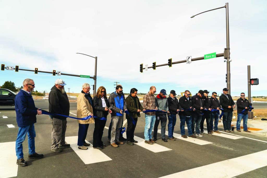 A group of people stand on a crosswalk cutting a blue ribbon at an intersection with traffic lights.