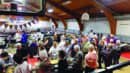 A large group of people standing in line at a buffet inside a gymnasium with wooden ceilings and basketball hoops.