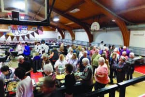 A large group of people standing in line at a buffet inside a gymnasium with wooden ceilings and basketball hoops.