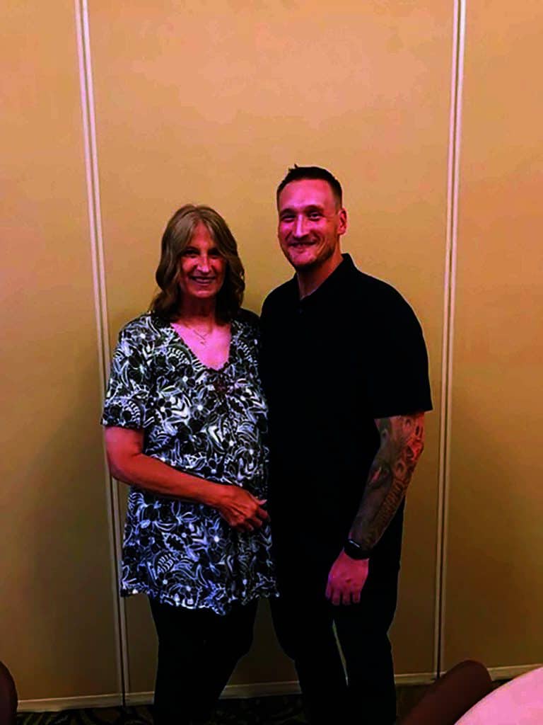 A woman in a patterned shirt and a man in a black polo stand side by side in a room with beige walls.