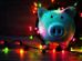 A white piggy bank wrapped in colorful string lights on a wooden surface.