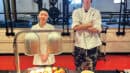 Two people in chef outfits stand behind a table with rolls, butter, and Hershey's syrup, in a gym with weightlifting equipment.