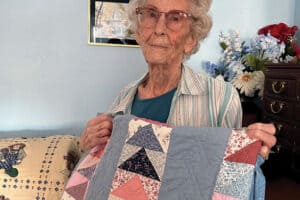 Elderly woman holding a colorful patchwork quilt in a room with framed pictures and flowers.