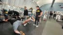 A person balances on one knee with a leg extended behind, while three others in casual sports attire observe in a gym setting.