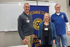 Three people stand in front of an Exchange Club banner in a classroom.