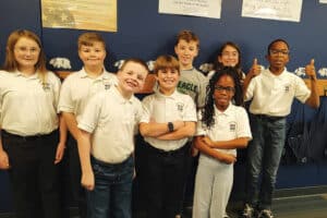 Eight children in matching white polo shirts and jeans stand in a classroom against a blue wall. Some children give thumbs up; they all smile. Posters and bags are visible in the background.