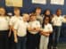 Eight children in matching white polo shirts and jeans stand in a classroom against a blue wall. Some children give thumbs up; they all smile. Posters and bags are visible in the background.