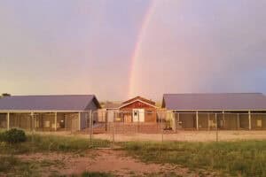 Two buildings with metal roofs sit behind a chain-link fence under a double rainbow against a cloudy sky.