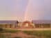 Two buildings with metal roofs sit behind a chain-link fence under a double rainbow against a cloudy sky.