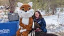 Person sitting next to a person in a fox costume, with a dog nearby, at a snowy outdoor event.