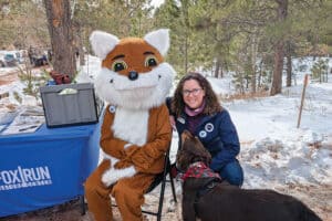 Person sitting next to a person in a fox costume, with a dog nearby, at a snowy outdoor event.