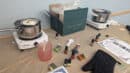 Candle-making setup with melting pots, fragrance bottles, wax pellets, and a green box on a table. Instructions and a black oven mitt are visible.