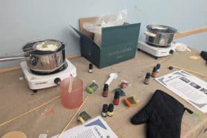 Candle-making setup with melting pots, fragrance bottles, wax pellets, and a green box on a table. Instructions and a black oven mitt are visible.