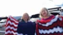 Two women standing in front of parked cars, each holding a crocheted blanket with red, white, and blue zigzag patterns.