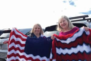 Two women standing in front of parked cars, each holding a crocheted blanket with red, white, and blue zigzag patterns.