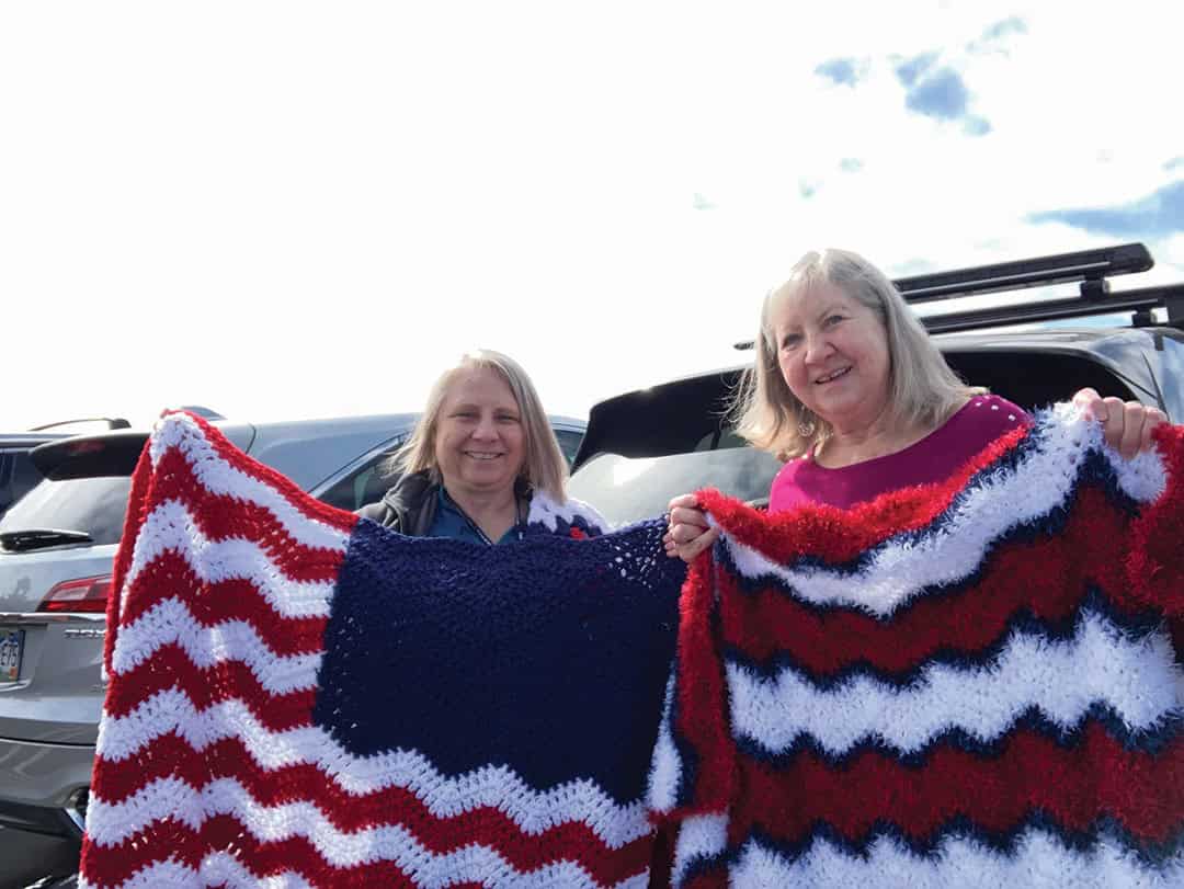 Two women standing in front of parked cars, each holding a crocheted blanket with red, white, and blue zigzag patterns.