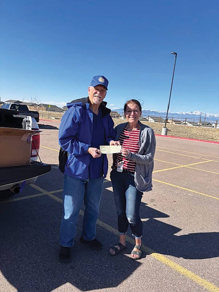 A man in a blue jacket and cap hands a check to a woman in a gray jacket. They stand in a parking lot with a clear sky and mountains in the background.