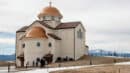 A beige church with copper domes stands on a slight hill, surrounded by snow patches. A few people are gathered near the building. Mountains are visible in the background under a cloudy sky.