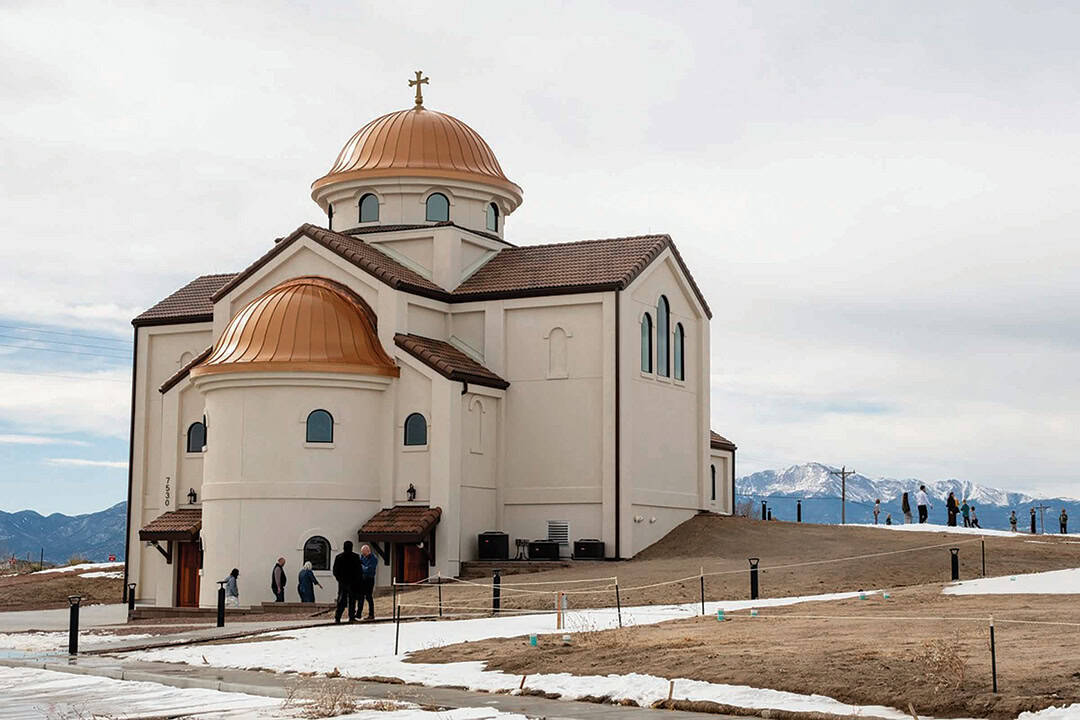 A beige church with copper domes stands on a slight hill, surrounded by snow patches. A few people are gathered near the building. Mountains are visible in the background under a cloudy sky.