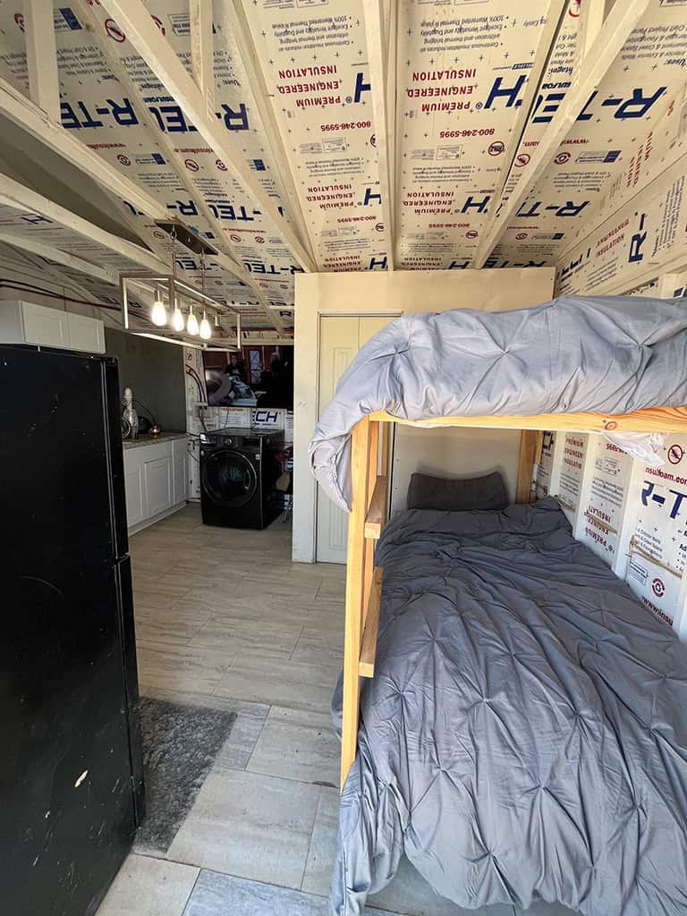 A small room with exposed insulation, a bunk bed with gray bedding, a black refrigerator, visible washer and dryer, and a kitchenette with white cabinets.