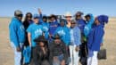 A group of people pose together outdoors on a sunny day, most wearing blue shirts and hoodies with a white logo, standing on dry grassland.