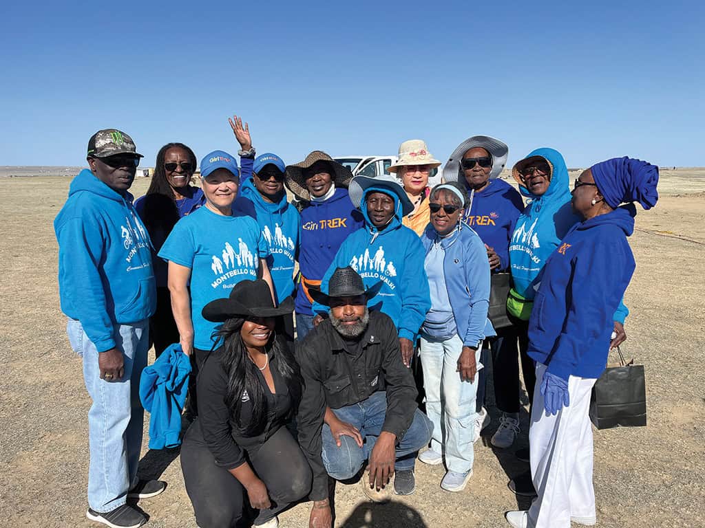 A group of people pose together outdoors on a sunny day, most wearing blue shirts and hoodies with a white logo, standing on dry grassland.