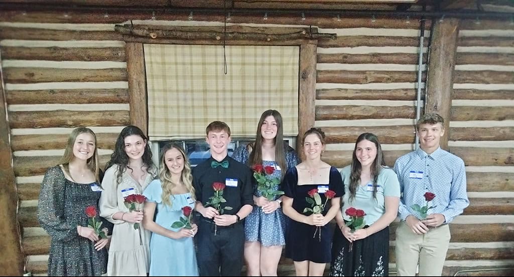 Eight young people stand in a row in front of a log cabin wall, each holding a red rose and wearing name tags.