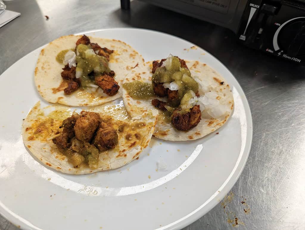 Three small tacos with grilled meat, chopped onions, and green salsa are arranged on a white plate on a metal surface.