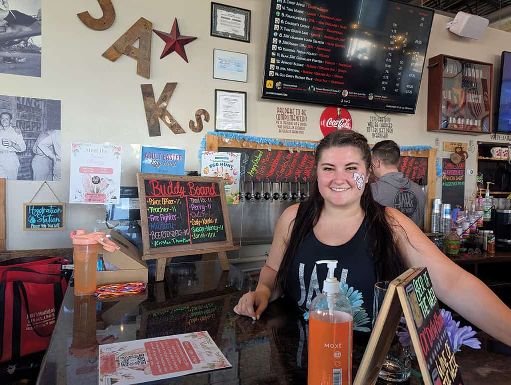 A woman stands and smiles behind a bar counter with beverage taps, drink menus, and informational signs visible in the background.