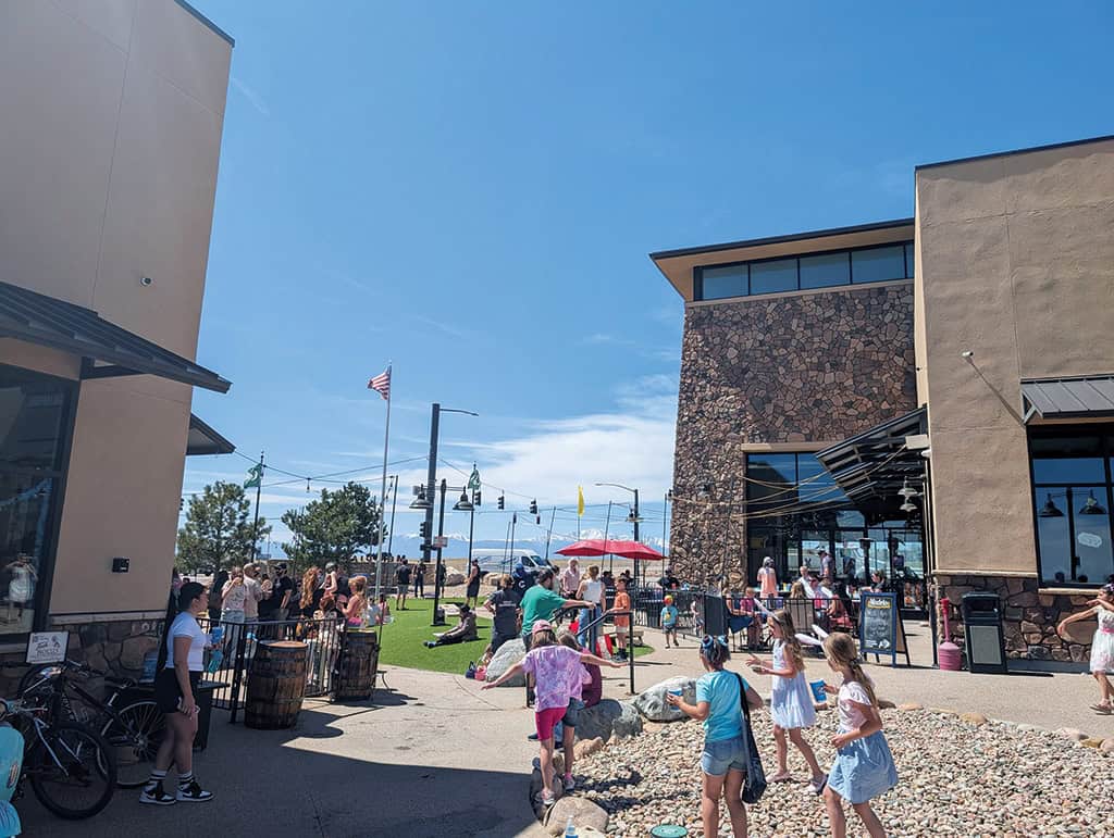 Children and adults gather in an outdoor shopping area on a sunny day, with people playing on grass and walking among shops.