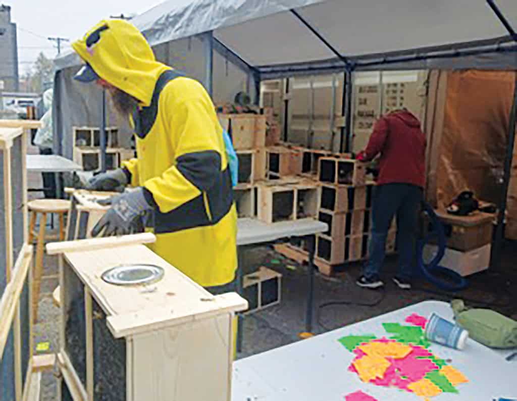 Two people assemble and sand wooden boxes under a tent; one wears a yellow hoodie, while colorful note papers and tools are visible on a table in the foreground.