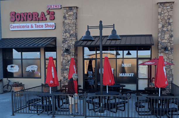 Outdoor seating area with red umbrellas in front of Sonora's Carnicería & Taco Shop, a meat market and restaurant in a beige building.