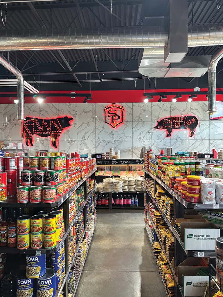 A grocery store aisle with shelves of canned goods and packaged foods, featuring neon butcher-style pig signs on a tiled wall above the shelves.