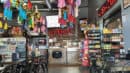 A brightly lit restaurant interior with colorful hanging decorations, metal chairs and tables, and shelves stocked with snacks and canned goods. The counter displays a sign reading "La Cocina.