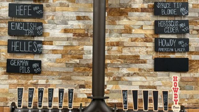 A row of twelve beer taps below a menu listing various beers on blackboards, mounted on a stone wall with glasses stored underneath.