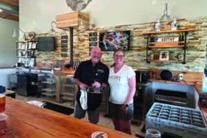 Two people stand behind a bar counter in a modern, well-lit bar with a brick accent wall, taps, framed photos, and a TV in the background.