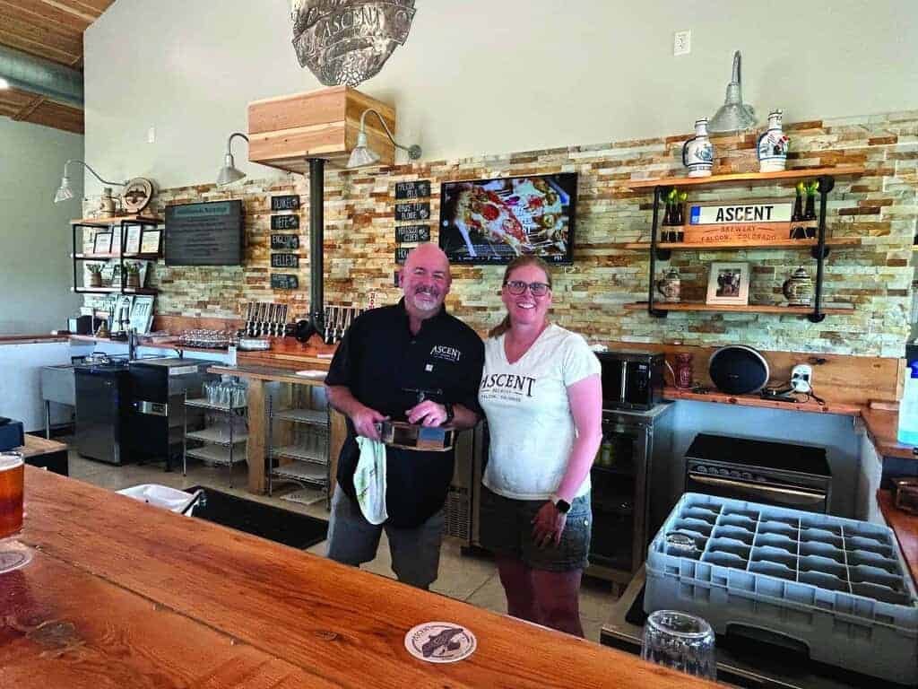 Two people stand behind a bar counter in a modern, well-lit bar with a brick accent wall, taps, framed photos, and a TV in the background.