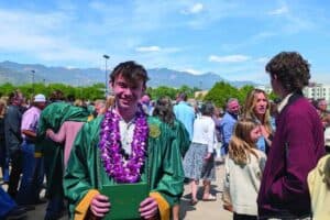 A graduate wearing a green gown and purple lei holds a diploma, standing among a crowd of people outdoors on a sunny day.