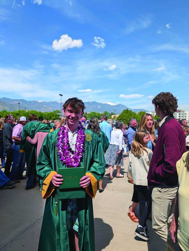 A graduate wearing a green gown and purple lei holds a diploma, standing among a crowd of people outdoors on a sunny day.