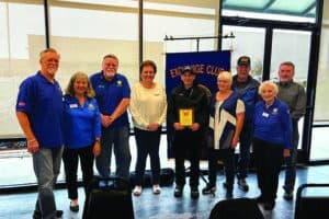 A group of nine people, some wearing blue shirts with name tags, stand indoors around a man holding a plaque in front of an "Exchange Club" banner.