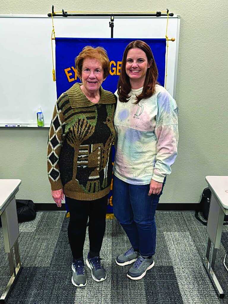 Two women stand side by side in a classroom setting, smiling at the camera. A blue and yellow banner and a whiteboard are visible in the background.