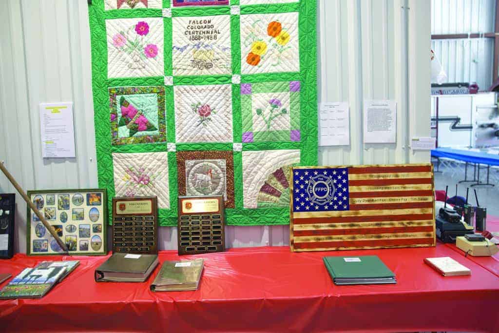 A display table with plaques, binders, and framed photos sits in front of a quilt and a wooden American flag with an emblem, all against a corrugated metal wall.
