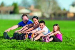 A family of four sits on grass in a park, stretching their legs forward and smiling at the camera on a sunny day.