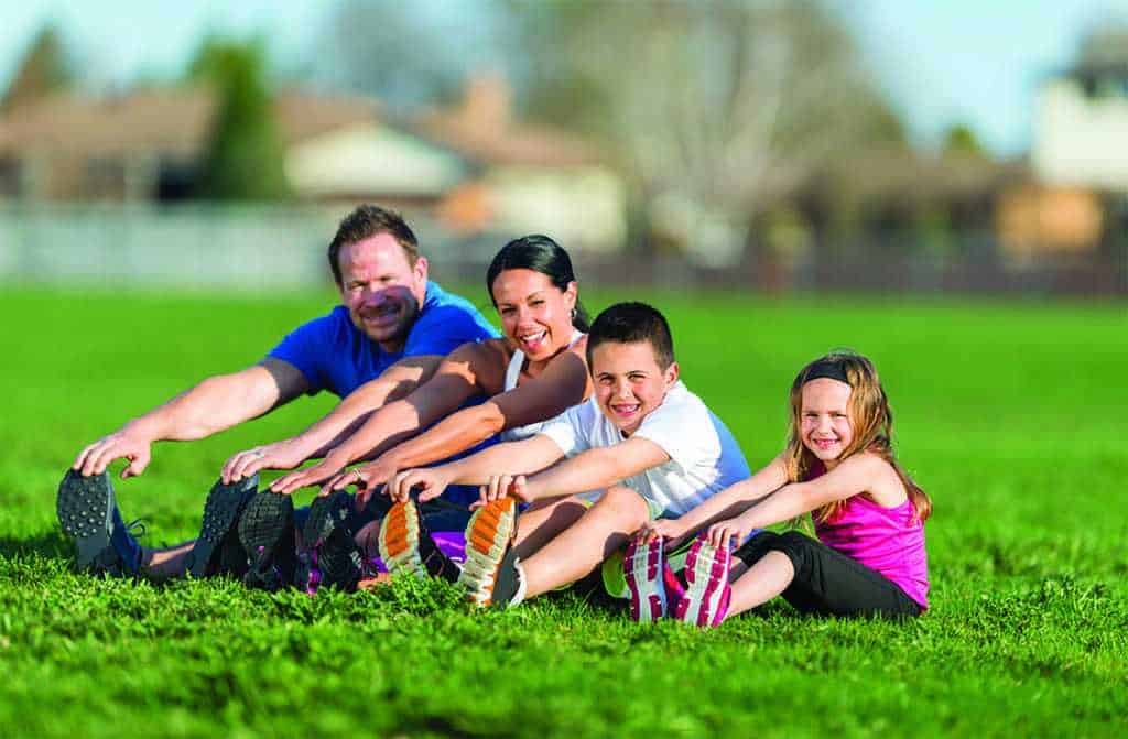 A family of four sits on grass in a park, stretching their legs forward and smiling at the camera on a sunny day.
