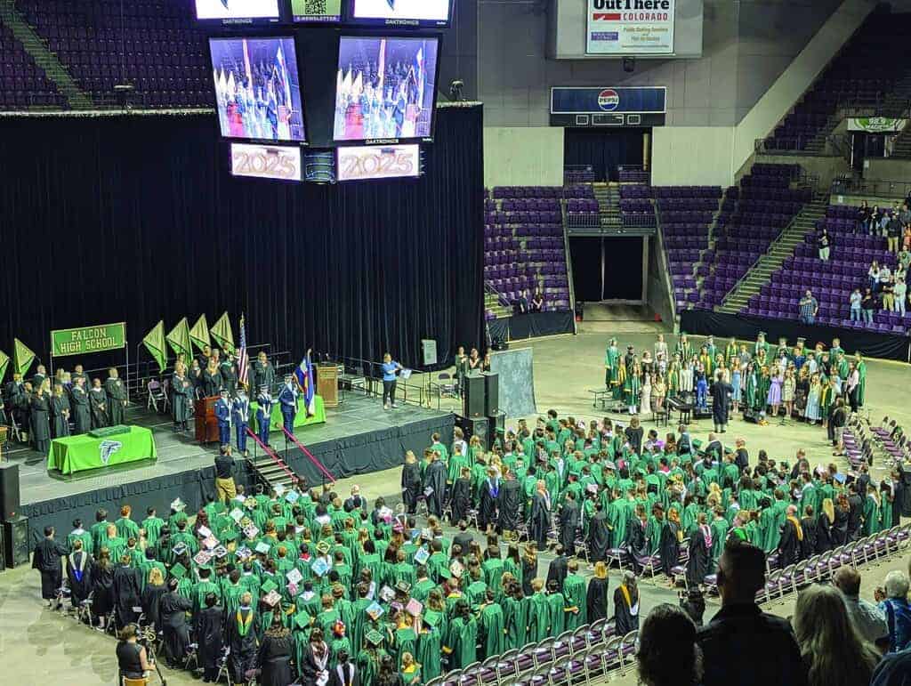 A large indoor graduation ceremony with rows of seated graduates in green gowns, an audience in purple seats, and a stage with speakers and banners.